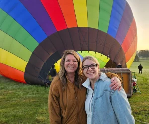 Mother and daughter smiling together in front of massive rainbow-colored hot air balloon at Rainbow Riders